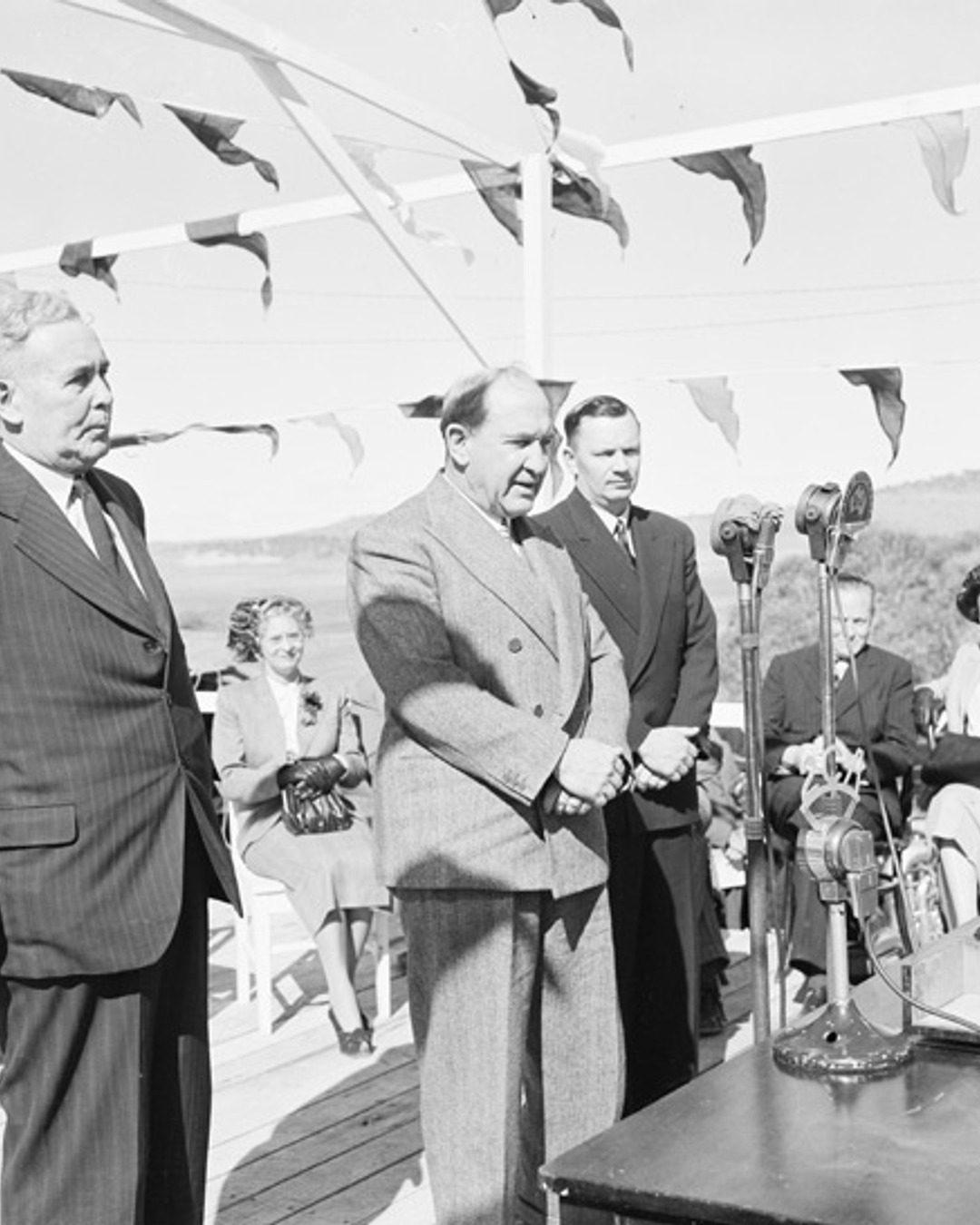 Black-and-white 1949 Department of Information photograph of officials at a microphone during an official Snowy Mountains Scheme launch event, with people gathered behind them.